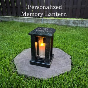 May include: A black granite memory lantern with a lit candle inside, set on a stone surface surrounded by green grass. The lantern has the text "Personalized Memory Lantern" and "Add Your Custom Text".