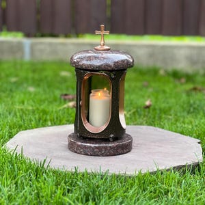 Może przedstawiać: Cylindryczny lampion nagrobny z granitu o polerowanym, brązowym nakrapianym wykończeniu, zwieńczony krzyżem w kolorze miedzi, z centralnym otworem zawierającym białą świecę pieńkową z ciepłym, płonącym płomieniem wewnątrz kamiennej konstrukcji.