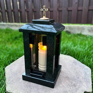May include: A black granite memorial lantern with a gold cross on top. The lantern has glass panels and a lit candle inside. The lantern is sitting on a stone surface, with a green grass background.