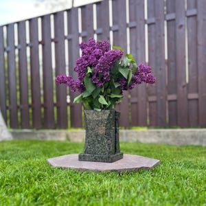 Könnte beinhalten: Eine Steinvase mit dunkelgrüner, gesprenkelter Oberfläche hält einen Strauß leuchtend violetter Flieder. Die Vase steht auf einem sechseckigen Steinsockel, mit einem Holzzaun im Hintergrund.