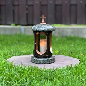 Puede incluir: Una linterna conmemorativa negra y bronce con una vela encendida en su interior. La linterna tiene una cruz en la parte superior y se apoya sobre una base de piedra. Fabricada en granito y bronce, para uso en exteriores.