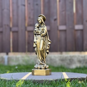 Może przedstawiać: Złota statuetka przedstawiająca postać w długiej, powłóczystej szacie trzymającą małe dziecko. Postać ma na głowie nakrycie i aureolę, stoi na prostokątnej podstawie. Rzeźba charakteryzuje się szczegółowymi fałdami materiału i metalicznym wykończeniem.