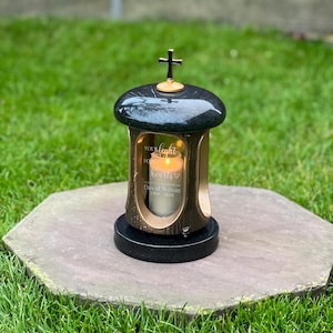 May include: A black and gold memorial lantern with a cross on top. The lantern features a glass panel with the words "Your light for hearts" and the name "David Wilson 1964-2024". A lit candle is visible inside.