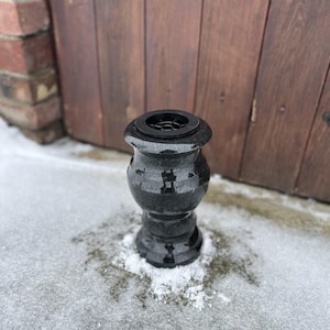 Puede incluir: Una urna de granito negro con forma decorativa, con base acampanada y parte superior redondeada con un inserto circular negro. La urna está colocada sobre una superficie nevada, con una puerta de madera y una pared de ladrillo al fondo.