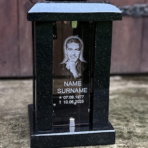 Könnte beinhalten: Eine Grablampe aus schwarzem Granit mit einer Glasscheibe, die ein eingraviertes Porträt, den Text NAME SURNAME sowie die Daten 07.09.1977 und 10.06.2025 zeigt. Die Struktur hat einen massiven Sockel und einen polierten Deckel.