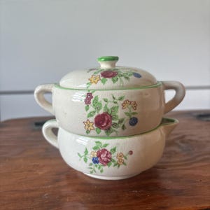 May include: Two-piece ceramic serving set. The set includes a lidded bowl and a smaller bowl, both with handles. The off-white pieces are decorated with a floral pattern featuring red roses, green leaves, and other colorful flowers. The rims are trimmed in green.