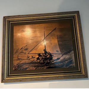 May include: A framed copper-toned artwork depicting a sailing vessel on a rail cart, with figures aboard and dogs running alongside. The artwork is set against a landscape with a tree and tall grass, framed in gold.