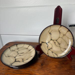 Puede incluir: Un juego de platos de cerámica beige y marrón con un patrón de grietas único. Los platos tienen un borde marrón oscuro y se exhiben sobre una superficie de madera. Un plato está sobre un soporte rojo. El juego de platos es decorativo.