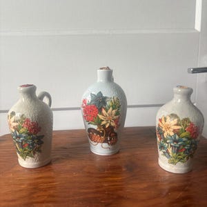 May include: Three ceramic vases with floral designs. The vases are light gray with a textured surface. The floral designs include red, yellow, and blue flowers. One vase has a handle. The vases are on a wooden surface.