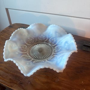 Puede incluir: Un cuenco de vidrio opalino con borde ondulado y borde festoneado. El cuenco presenta un diseño central texturizado rodeado de motivos florales. El vidrio tiene un color blanco lechoso y azul pálido, creando una apariencia suave y etérea. Este cuenco decorativo es ideal para servir o exhibir.