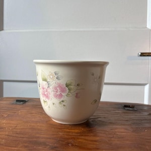 Pode incluir: Jarro de cerâmica creme com um design floral. O jarro apresenta flores rosa e brancas com folhas verdes pintadas no exterior. O jarro está sobre uma superfície de madeira. É um artigo decorativo.