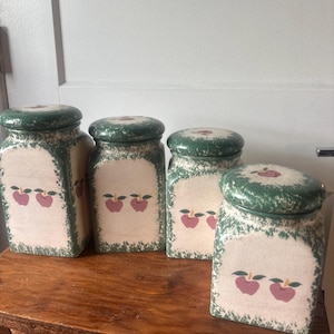 Può includere: Quattro barattoli in ceramica con coperchi verdi e corpo color crema. Ogni barattolo presenta un disegno dipinto di due mele rosse con foglie verdi. I coperchi e i bordi hanno un motivo verde maculato. I barattoli sono esposti su una superficie di legno.