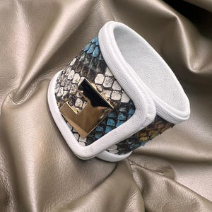Puede incluir: Un brazalete de cuero blanco con un patrón de piel de serpiente y un acento de metal dorado en forma de letra 'F'.