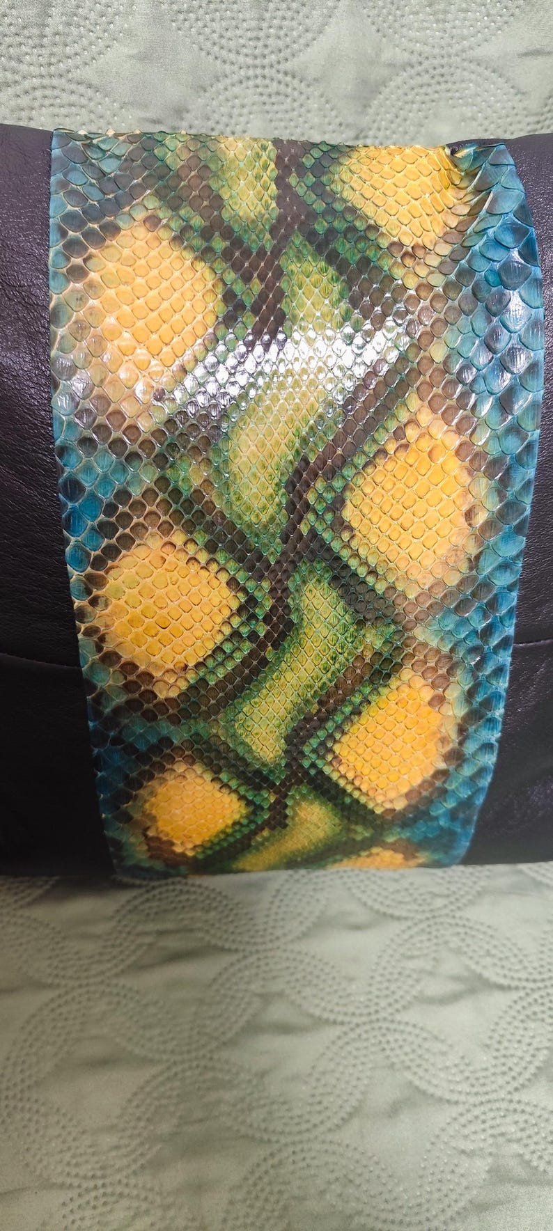 Op de afbeelding: Een decoratief kussen met een zwarte leren achterkant en een voorpaneel van gele, groene en blauwe slangenleer.