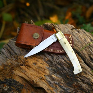 Puede incluir: Un cuchillo plegable con una hoja de acero pulido y un mango de hueso descansa sobre una superficie de madera envejecida. El cuchillo est&aacute; acompa&ntilde;ado de una funda de cuero marr&oacute;n con cierre de bot&oacute;n decorativo. La hoja est&aacute; abierta, mostrando su filo.