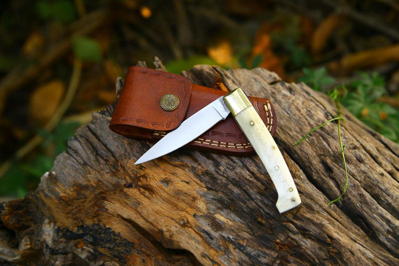 Puede incluir: Un cuchillo plegable con una hoja de acero pulido y un mango de hueso descansa sobre una superficie de madera envejecida. El cuchillo est&aacute; acompa&ntilde;ado de una funda de cuero marr&oacute;n con cierre de bot&oacute;n decorativo. La hoja est&aacute; abierta, mostrando su filo.