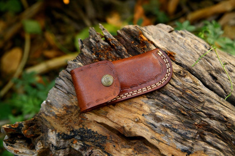 Puede incluir: Una funda de cuero marr&oacute;n para cuchillo con cierre de bot&oacute;n decorativo y costuras blancas. La funda descansa sobre un trozo de madera desgastada, con un fondo borroso de follaje verde.