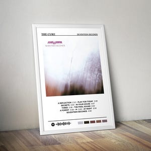 Puede incluir: Un póster enmarcado en blanco con una foto en blanco y negro de un bosque borroso. El póster tiene el título "The Cure" y el título del álbum "Seventeen Seconds" en texto negro. La lista de canciones del álbum se muestra debajo de la foto con los títulos de las canciones y las duraciones en texto negro.