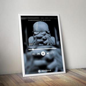 May include: A framed black and white print featuring the album art for "Breaking Benjamin We Are Not Alone." The image shows a person with their hands clasped over their face, with a Spotify-style music player interface overlayed.