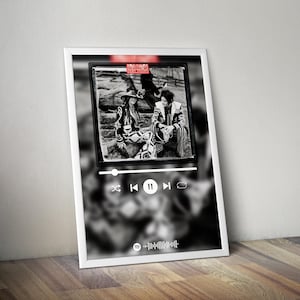 May include: A framed black and white print featuring a photograph of two people in patterned outfits. The print is designed to look like a music player interface, with play/pause and skip controls. The frame is white, and the print is leaning against a wooden surface.