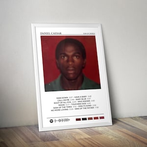 May include: A framed poster featuring a portrait of Daniel Caesar with the text "SON OF SPERGY." The poster includes a tracklist with song titles and durations. The color scheme is primarily red, white, and black.