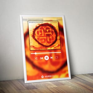May include: A framed print featuring the album art for "Saturate" by Breaking Benjamin. The artwork is a circular design in shades of orange and red. The print includes a music player interface with play, pause, and skip controls.