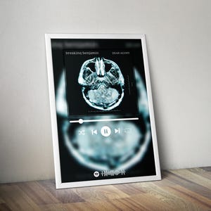 May include: A framed print featuring a digital art design. The artwork displays a stylized brain scan image with the text "breaking benjamin" and "DEAR AGONY". Below the image is a music player interface. The print is in a white frame, leaning against a wall.