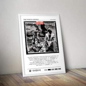 May include: A framed poster featuring the album art for "Icky Thump" by The White Stripes. The black and white image shows two people in patterned outfits. The album title and tracklist are displayed below the image. The poster is displayed on a wooden surface.