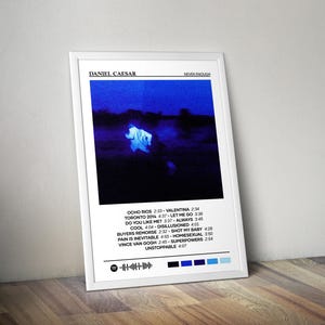 May include: A framed poster featuring the album art for Daniel Caesar's "Never Enough." The artwork has a blue and white color scheme. The poster includes the album's tracklist and a Spotify code at the bottom.