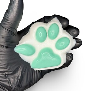 Könnte beinhalten: Eine pfotenförmige Seife mit weißer Basis und mintgrünen Pads. Die Seife wird von einer behandschuhten Hand vor weißem Hintergrund gehalten. Das Pfotenabdruck-Design ist ein beliebtes Thema für Tierliebhaber.