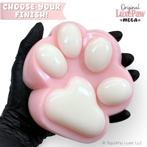 以下が含まれることがあります： CHOOSE YOUR FINISH!およびOriginal LuxePaw MEGAというテキストが記載された、ピンクと白の肉球型オブジェクト。滑らかで丸みを帯びたピンクのベースに、4つの隆起した光沢のある白い楕円形のパッドと、中央に大きな白いパッドが配置されています。