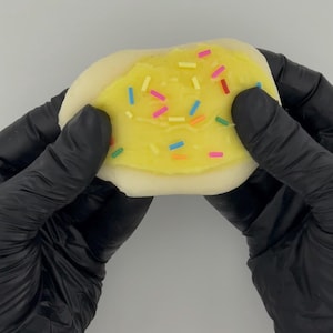 May include: A white and yellow cookie with yellow frosting and colorful sprinkles. The cookie is being held by two gloved hands.