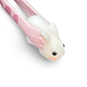May include: Pink and white bunny-shaped kitchen tongs with pink polka dots. The tongs are holding a white bunny-shaped object.