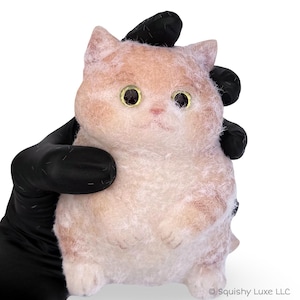 May include: A small, fuzzy, peach-colored cat figurine with large, round, dark eyes and a light-colored, textured surface. The cat has pointed ears, a small nose, and a rounded body shape. The text Squishy Luxe LLC is visible in the bottom corner.