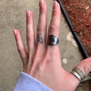 May include: A person's hand wearing two silver rings. The ring on the index finger is a simple band, while the ring on the middle finger is a wide band with a textured design. The person also has a small butterfly tattoo on their finger.