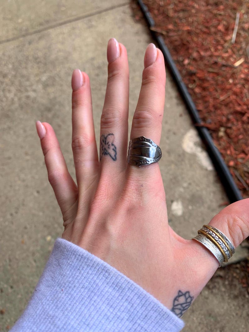 May include: A person's hand wearing two silver rings. The ring on the index finger is a simple band, while the ring on the middle finger is a wide band with a textured design. The person also has a small butterfly tattoo on their finger.
