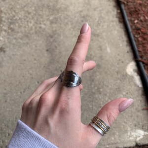 May include: A silver spoon ring with a decorative design on the top. The ring is worn on the index finger of a hand. A stack of three silver and gold rings is worn on the thumb.