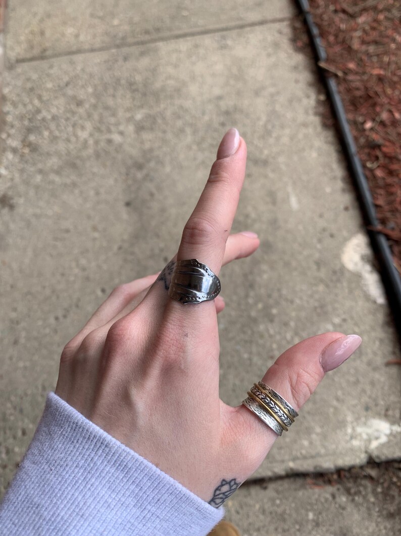 May include: A silver spoon ring with a decorative design on the top. The ring is worn on the index finger of a hand. A stack of three silver and gold rings is worn on the thumb.
