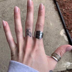 May include: A hand wearing a silver ring with a unique design and a silver band ring with a gold band. The hand also has a small butterfly tattoo on the finger.