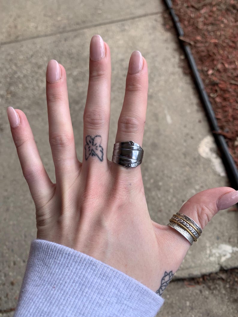 May include: A hand wearing a silver ring with a unique design and a silver band ring with a gold band. The hand also has a small butterfly tattoo on the finger.