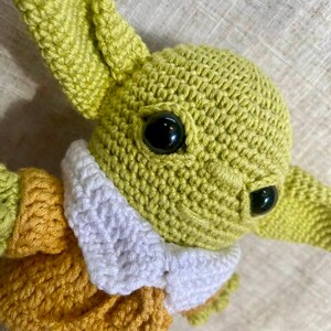 May include: A handmade crochet doll of a character with large black eyes and pointed ears. The doll is primarily light green with a white collar and a yellow and green outfit. The close-up shot highlights the textured crochet stitches.