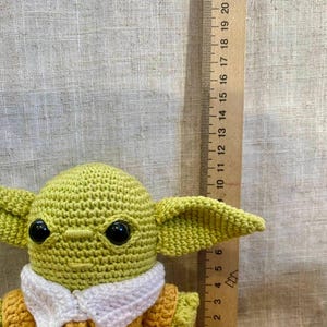 May include: A crocheted figure of a character with large ears and black eyes, wearing a yellow and white outfit. A wooden ruler with inch markings is next to the figure, showing its size.