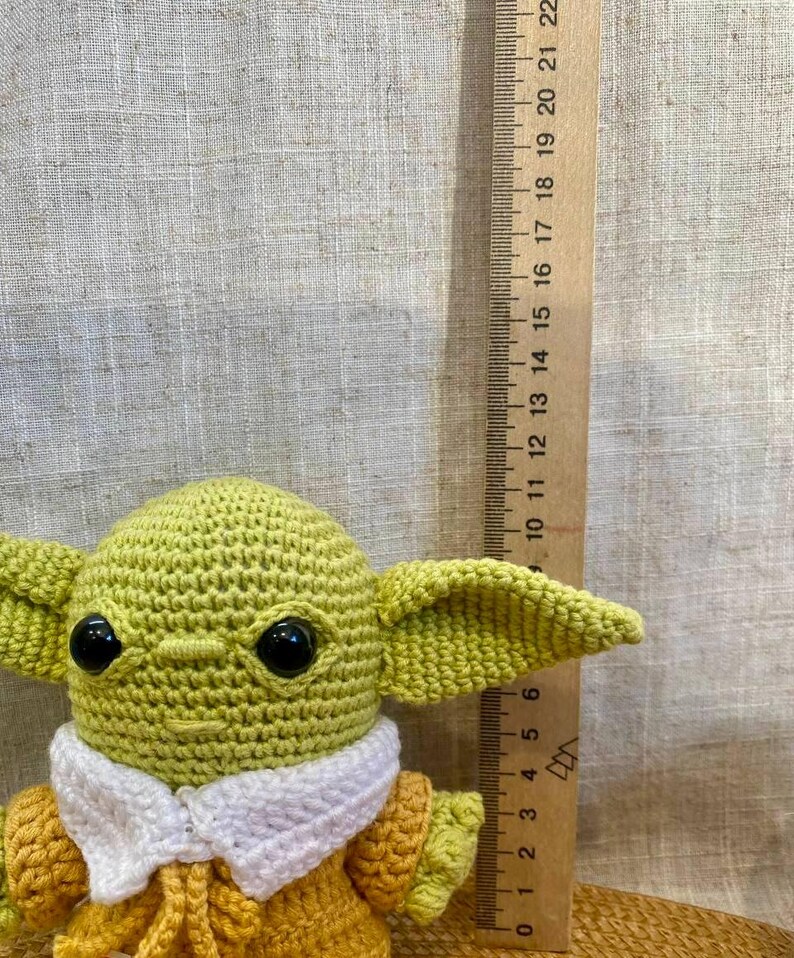 May include: A crocheted figure of a character with large ears and black eyes, wearing a yellow and white outfit. A wooden ruler with inch markings is next to the figure, showing its size.