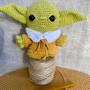May include: A handmade crochet figure of a character with large ears, black eyes, and a yellow-green complexion. The figure wears a yellow and white outfit. It is displayed on a spool of twine with a crochet hook.