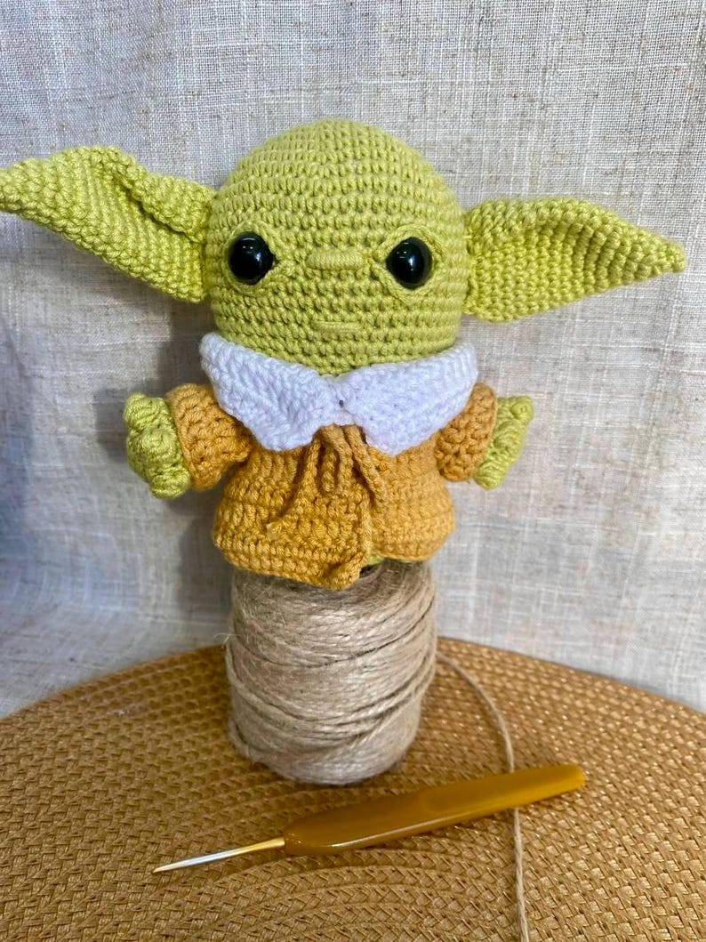 May include: A handmade crochet figure of a character with large ears, black eyes, and a yellow-green complexion. The figure wears a yellow and white outfit. It is displayed on a spool of twine with a crochet hook.