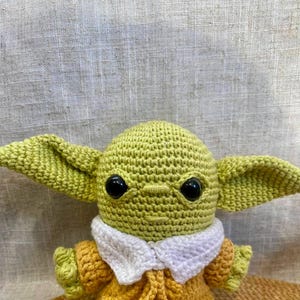 May include: A handmade crochet doll featuring a character with large ears, black eyes, and a yellow-green complexion. The doll wears a yellow and orange outfit with a white collar. The doll is approximately 6 inches tall.