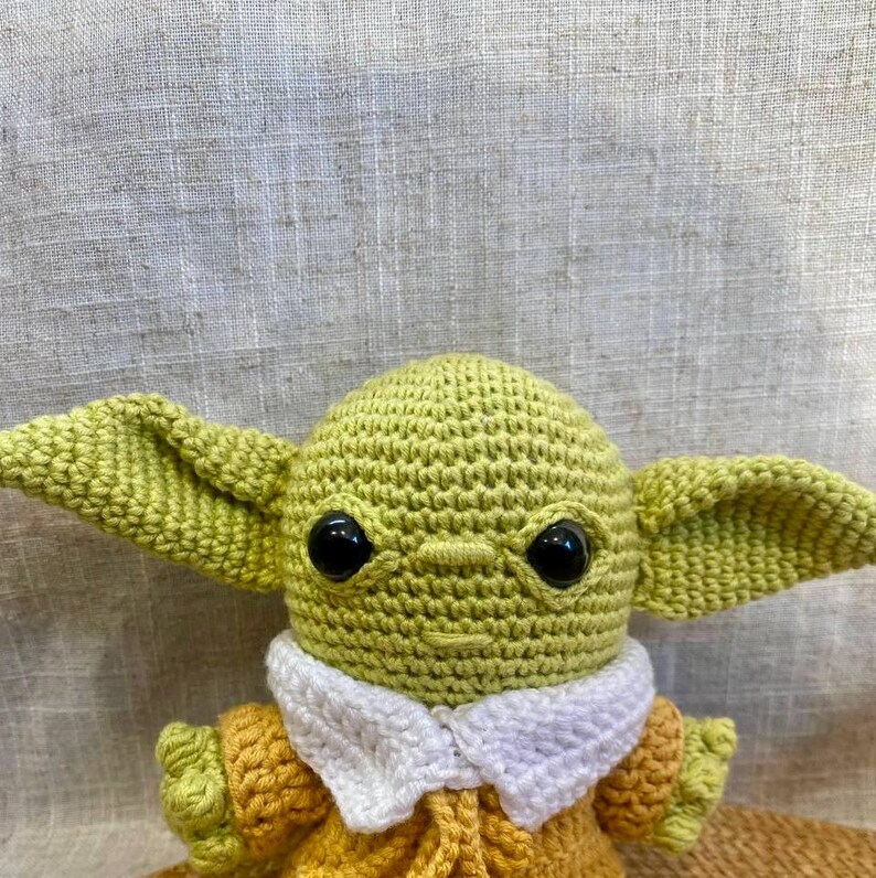 May include: A handmade crochet doll featuring a character with large ears, black eyes, and a yellow-green complexion. The doll wears a yellow and orange outfit with a white collar. The doll is approximately 6 inches tall.