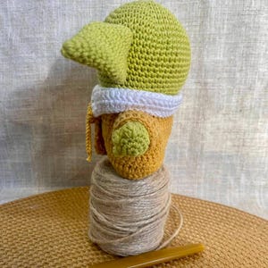 May include: A handmade crochet doll in the likeness of a character from a popular science fiction series. The doll features a green head with large ears, a yellow and white outfit, and is perched atop a spool of twine. A crochet hook rests nearby.