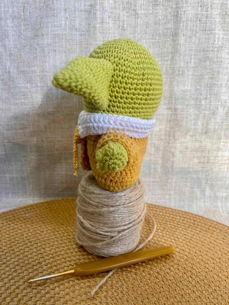 May include: A handmade crochet doll in the likeness of a character from a popular science fiction series. The doll features a green head with large ears, a yellow and white outfit, and is perched atop a spool of twine. A crochet hook rests nearby.