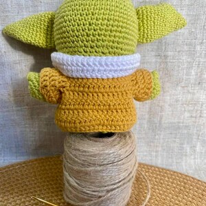 May include: A handmade crochet figure of a character with large ears, wearing a yellow and white outfit. The figure is perched atop a spool of twine, with a crochet hook in the foreground. The figure is approximately 4 inches tall.